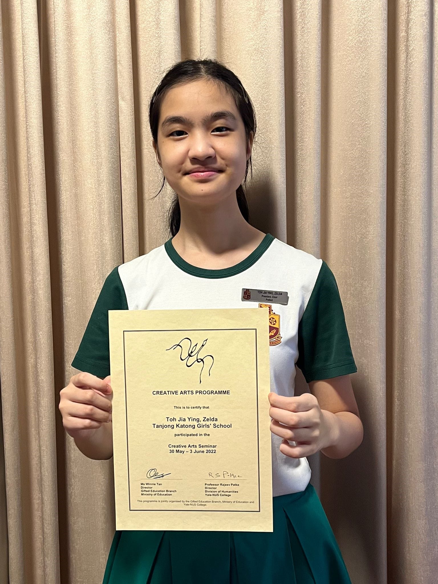 A young girl in a green and white school uniform holds a Creative Arts Programme certificate from June 2022.