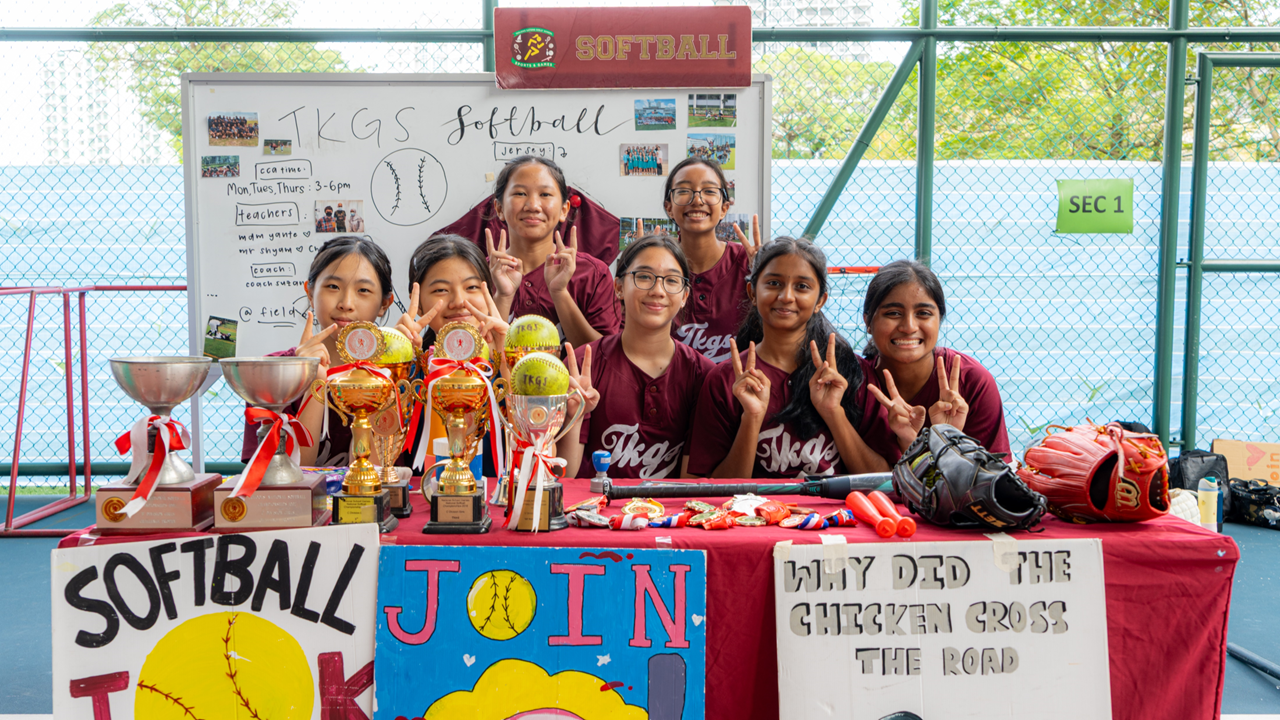 Seven girls in maroon TKGS softball uniforms pose behind a table displaying trophies, gloves, and recruitment posters.