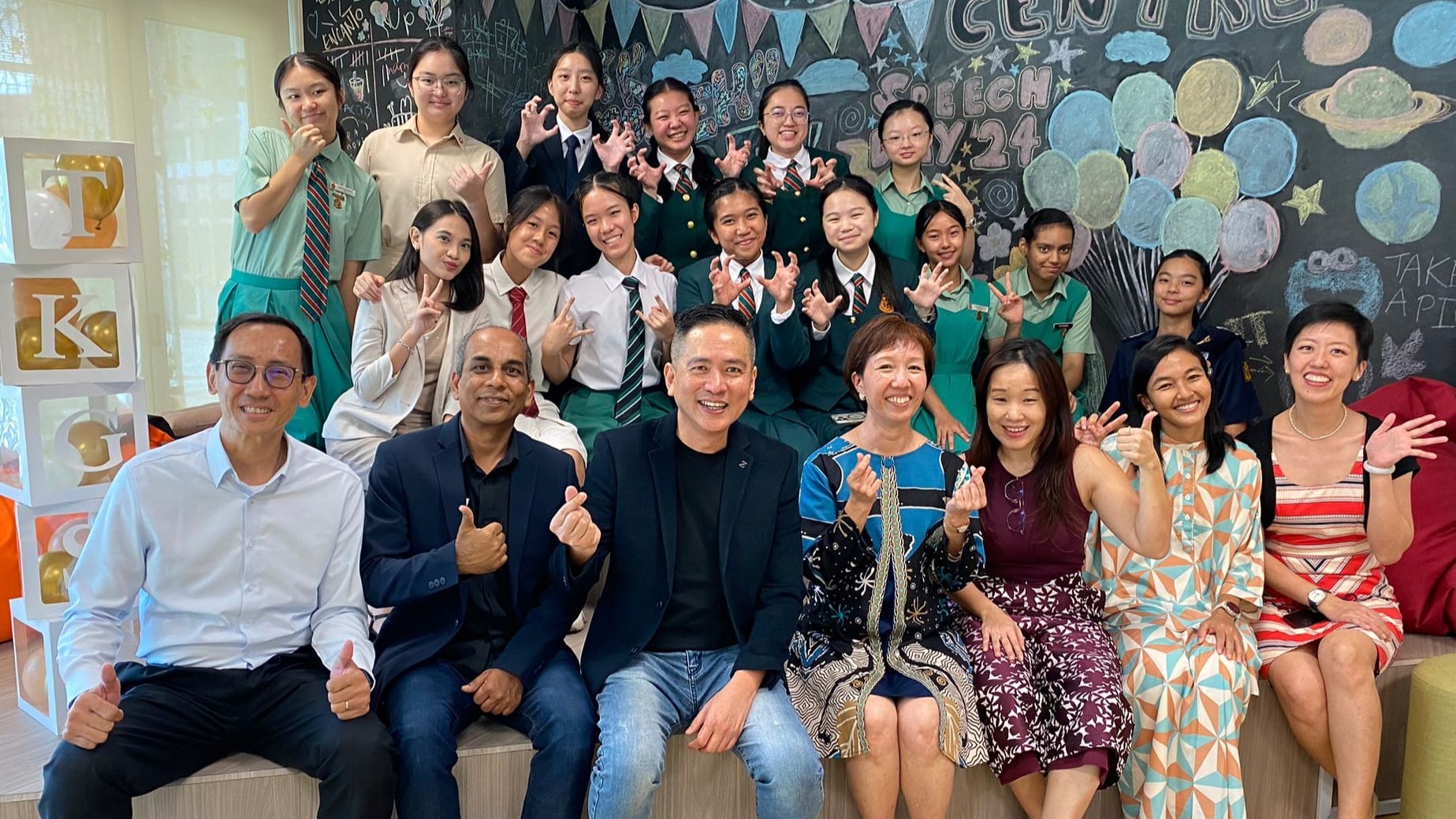 A large group of students in school uniforms and adults pose smiling before a decorated chalkboard reading "Speech Day 24."