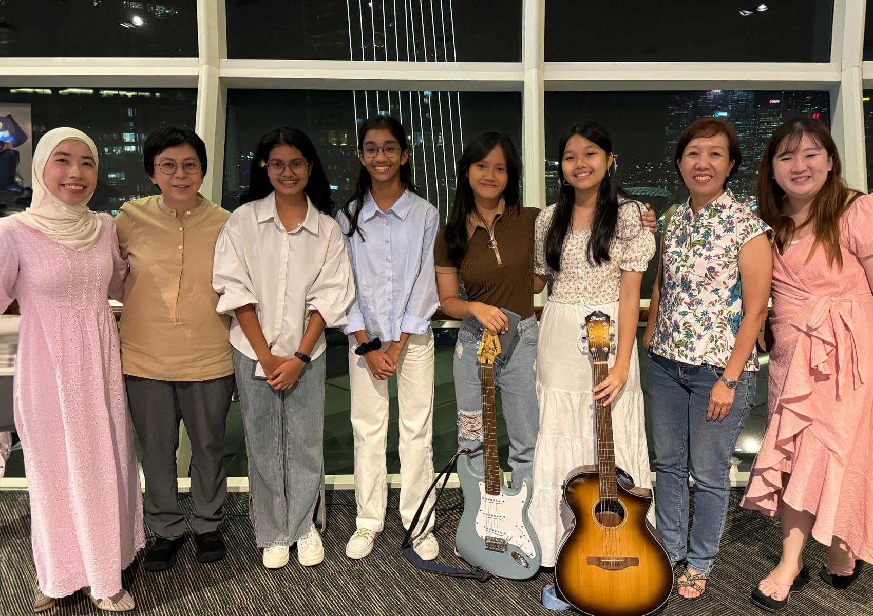 Eight women smiling together indoors at night, two holding guitars, with a city skyline visible through large windows.