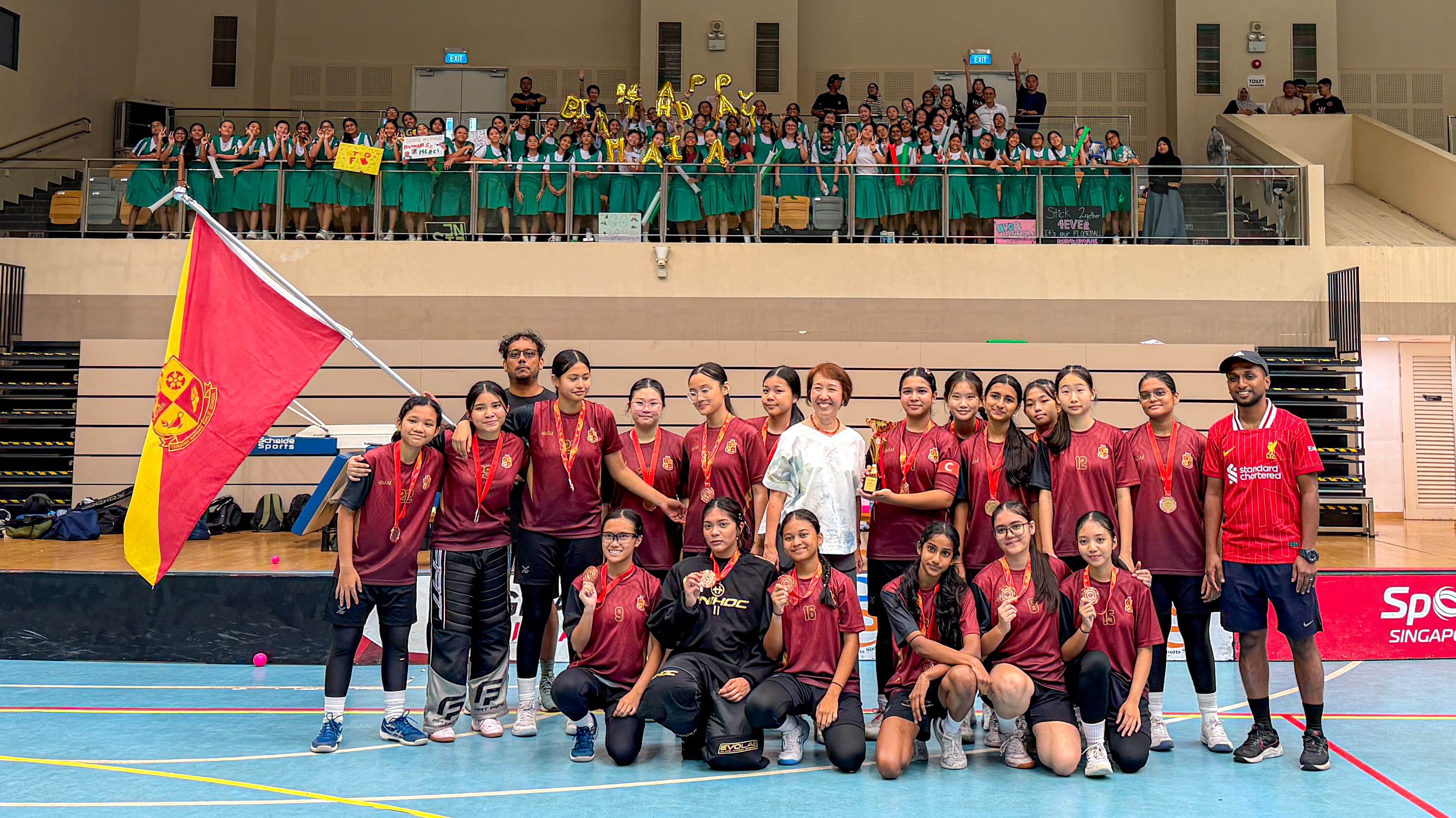 A girls sports team in maroon jerseys poses with medals, a trophy, and a red flag inside an indoor court.