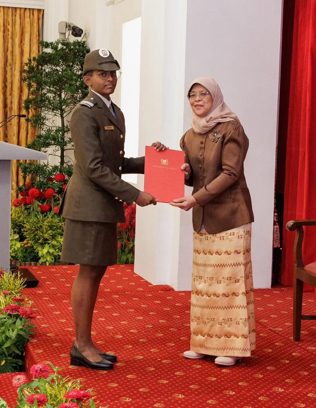 A female soldier in olive uniform receives a red certificate folder from a woman in a hijab at a formal ceremony.