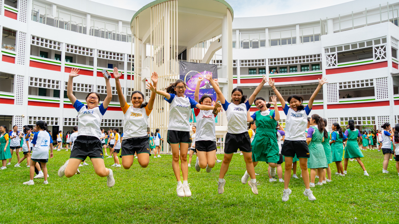 A group of school girls in uniforms jumping joyfully on a grass courtyard in front of a multi-story school building.