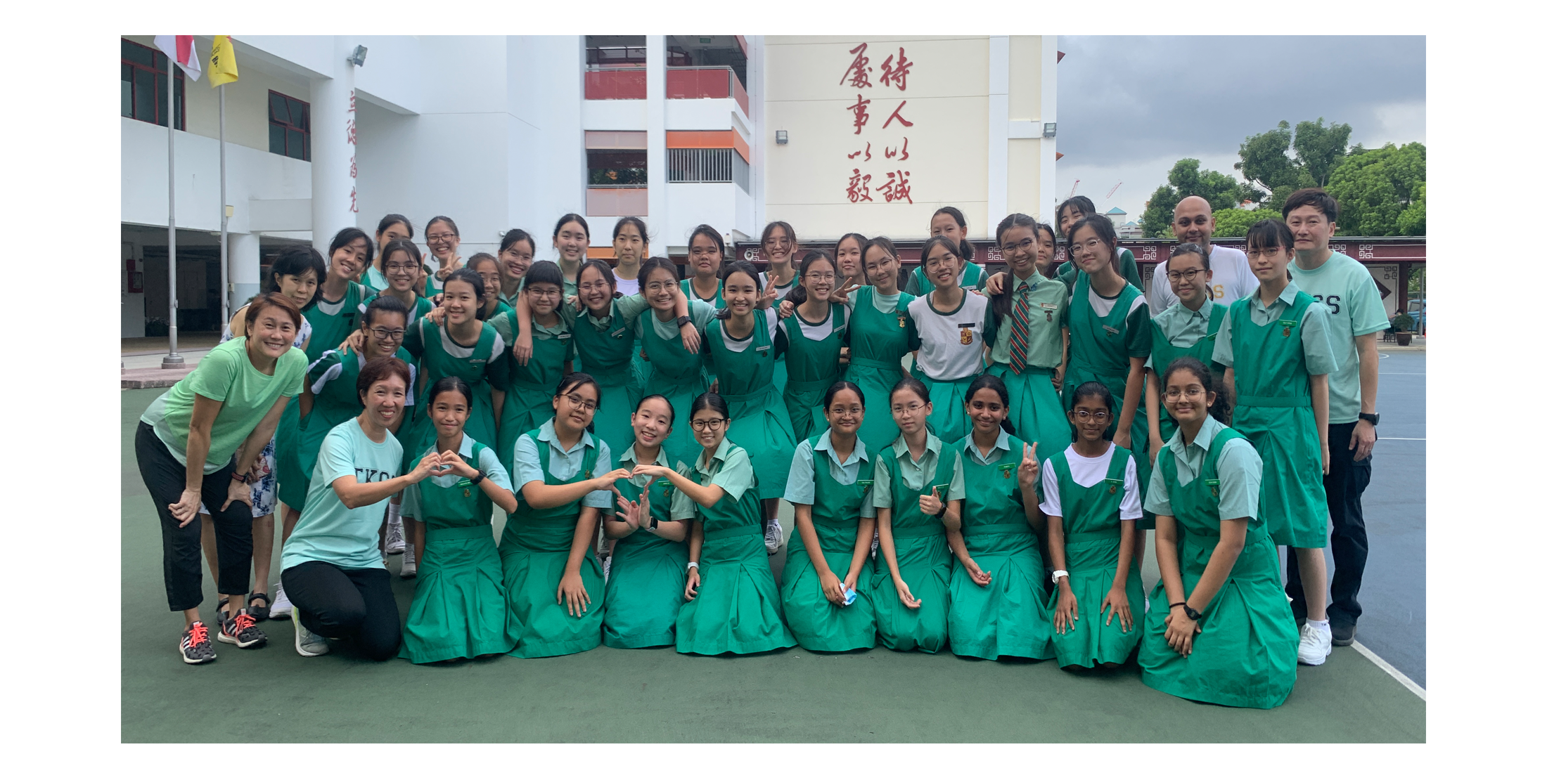 A large group of students in green uniforms pose with teachers outside a school building with Chinese characters.