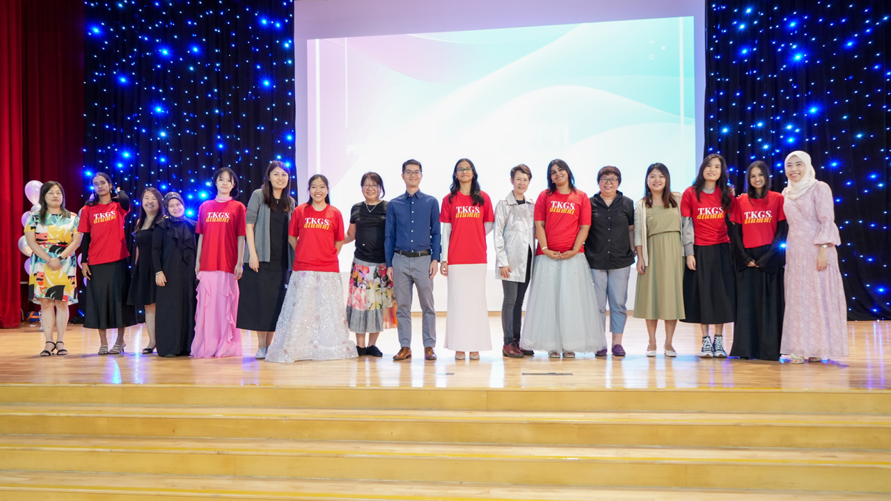 A group of people posing on a stage, several wearing red TKGS Alumni shirts, with a starlit backdrop.