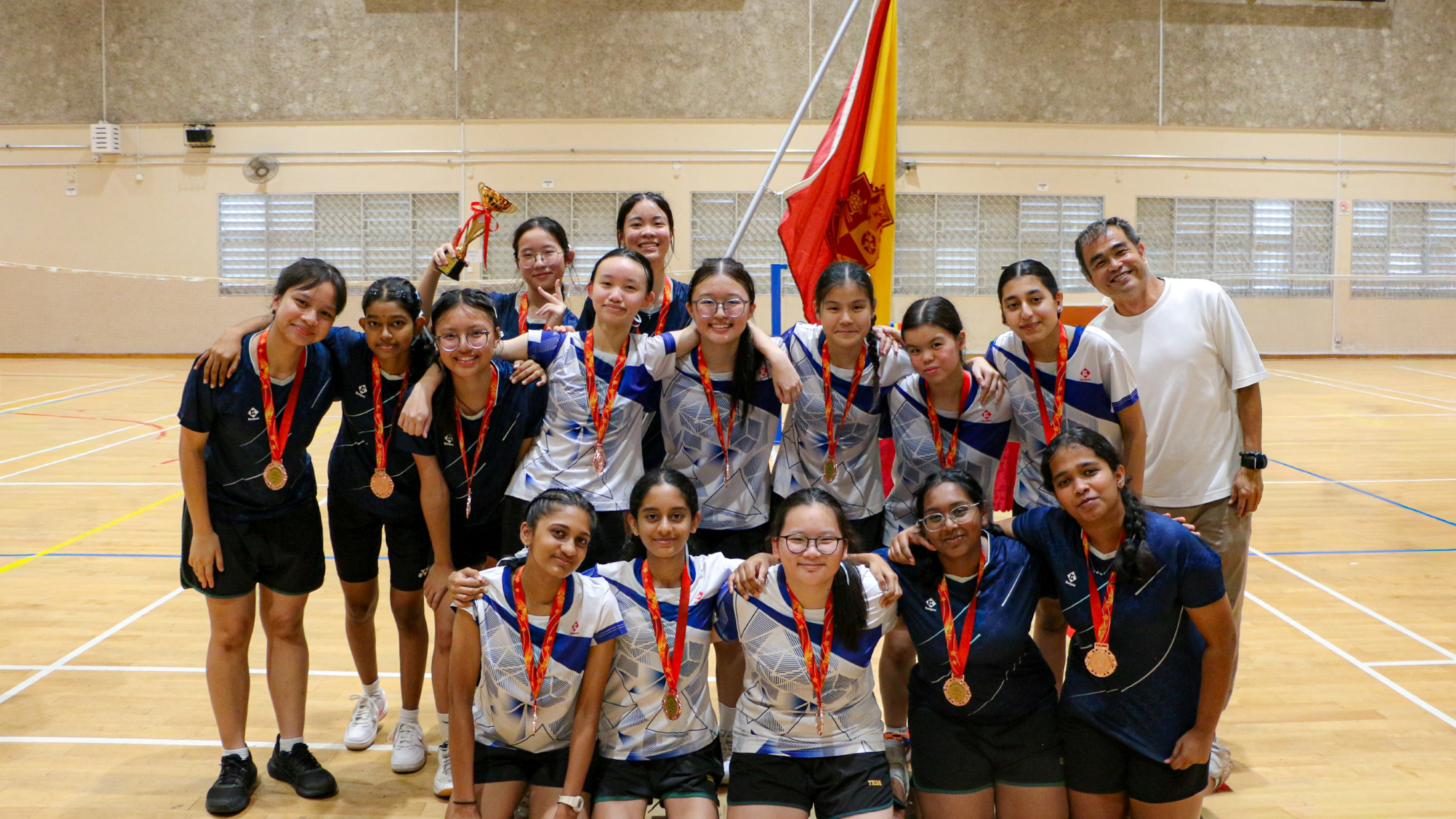 Girls’ volleyball team with medals poses in a gym, smiling beside coach and holding a red-and-yellow flag.