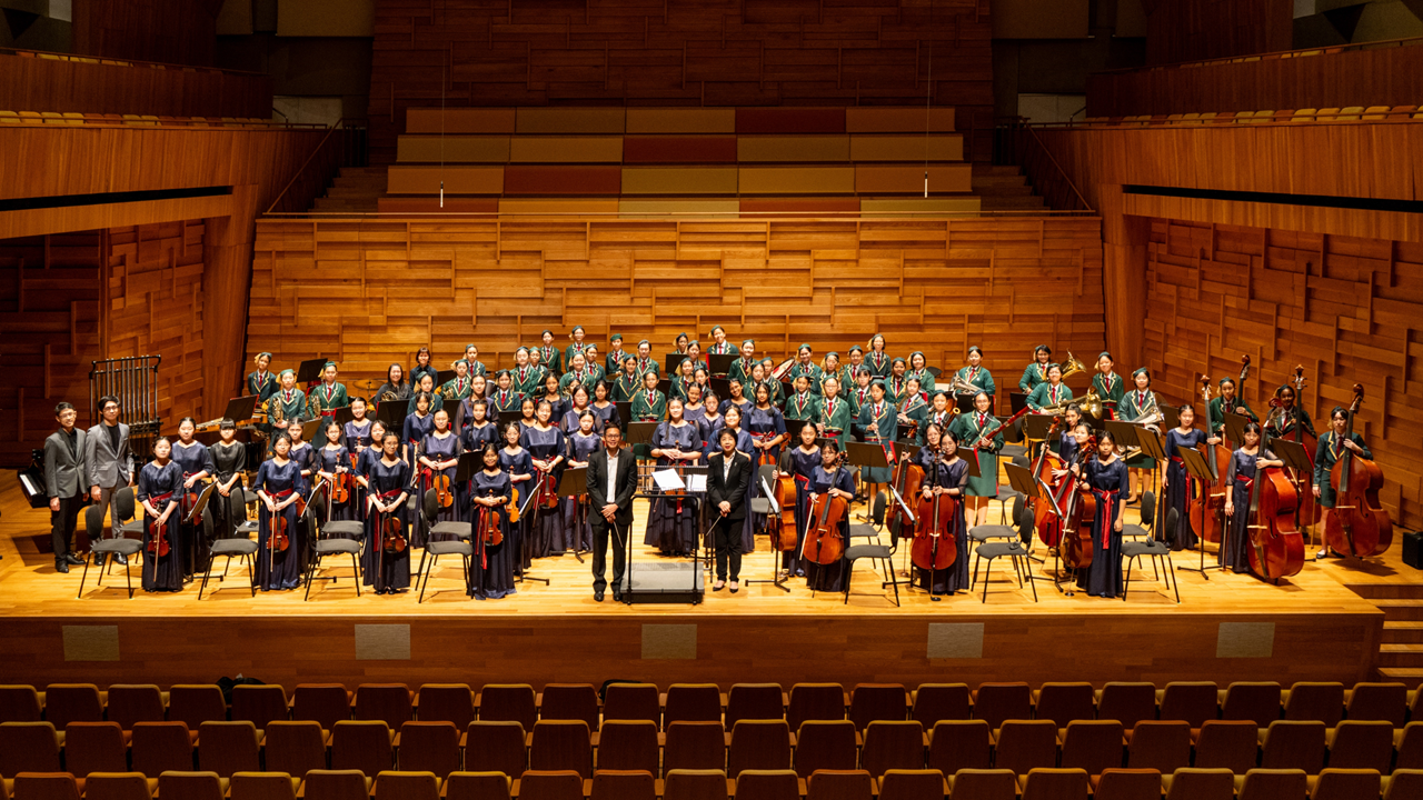 A large youth orchestra in formal uniforms poses on a wooden concert hall stage with a conductor at center.