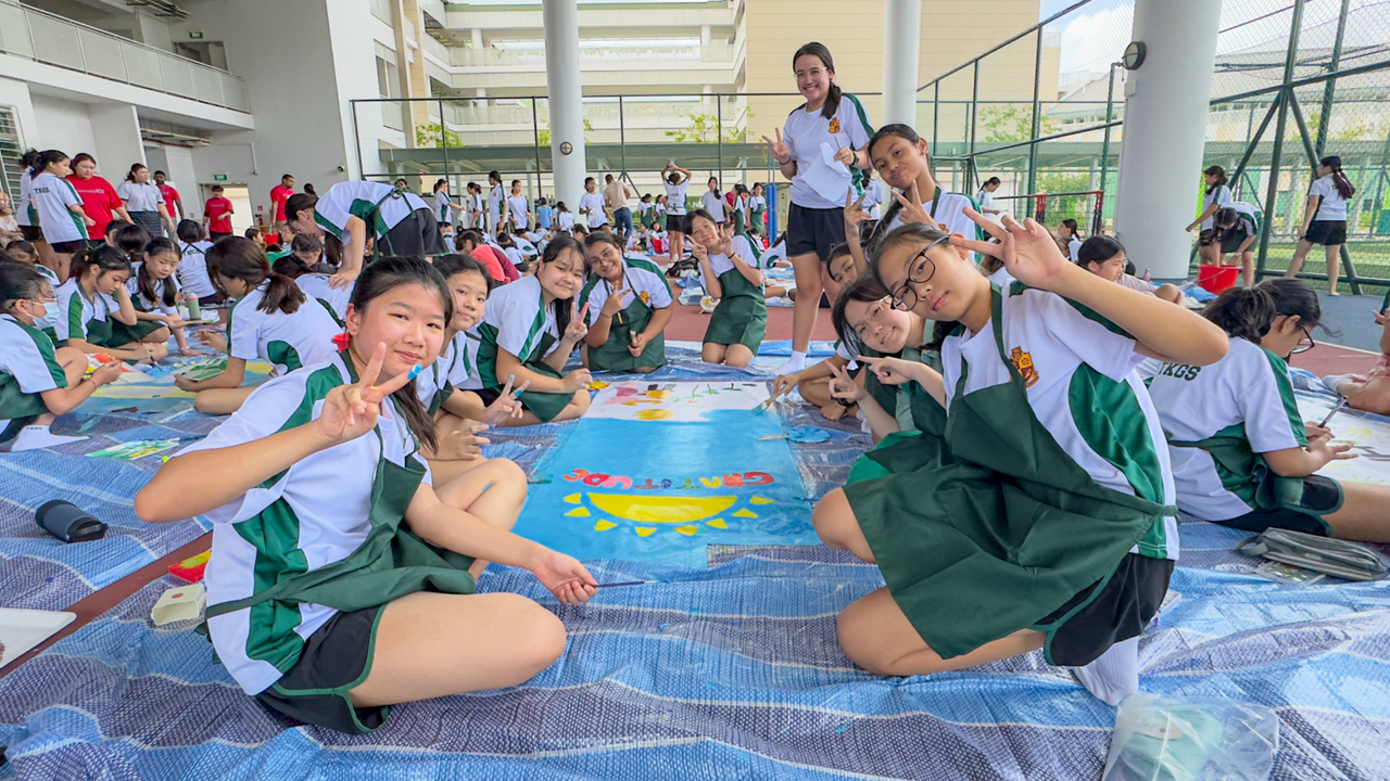 Students in white and green uniforms sit on a courtyard floor, painting a large colorful mural and posing with peace signs.