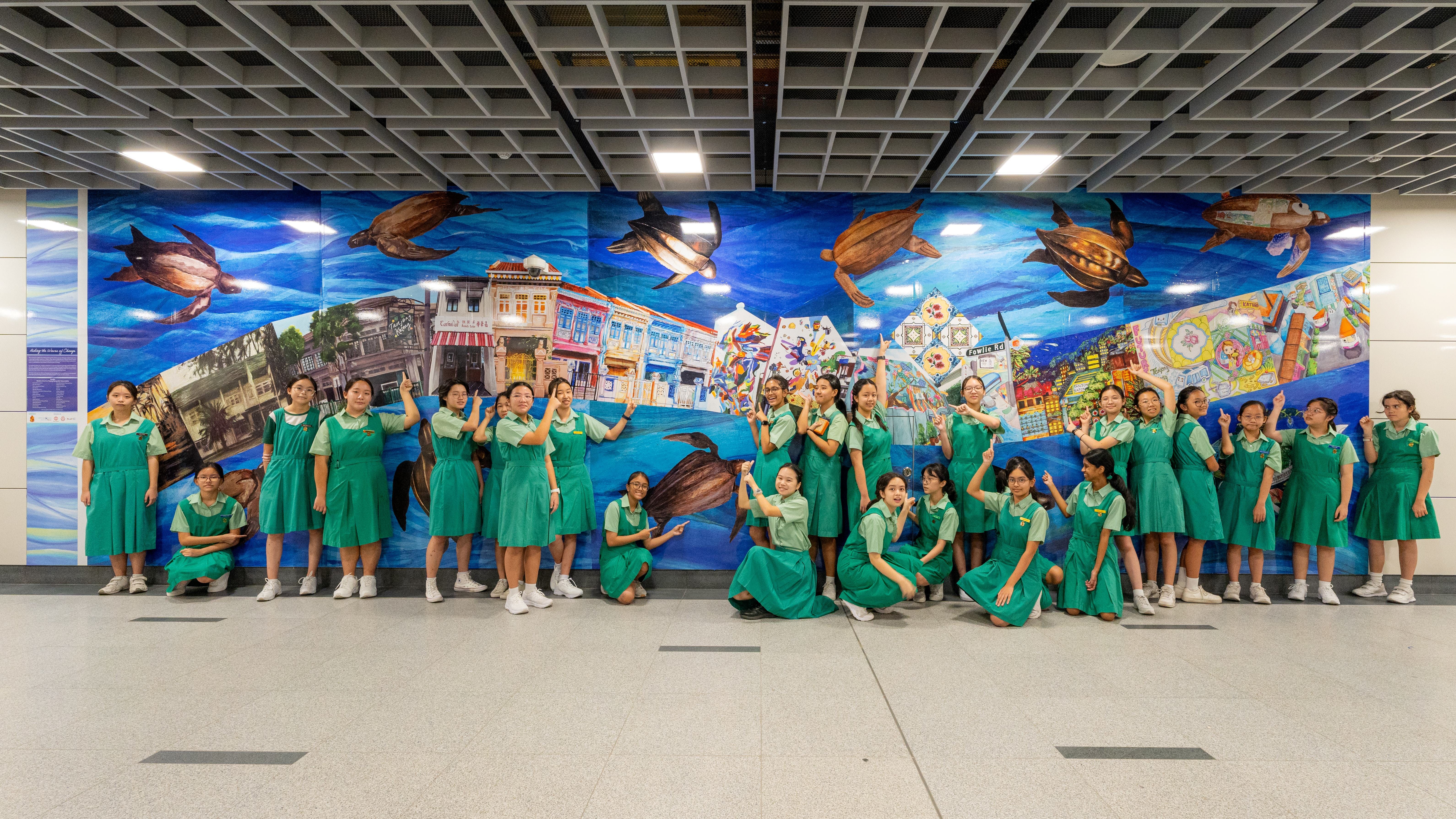 Group in green school uniforms pose in front of a bright mural with sea turtles and city scenes.