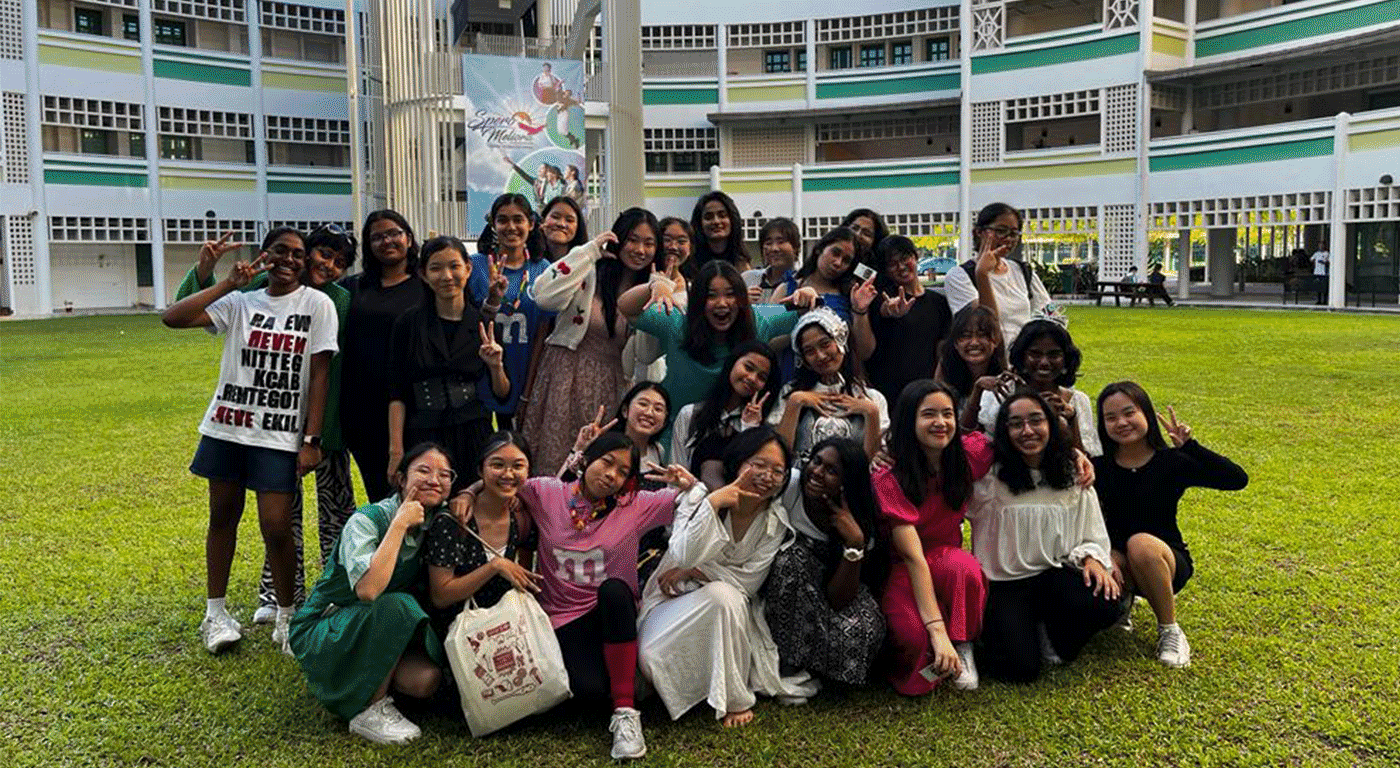 A large group of young women pose together on a green lawn in front of a multi-story school building.