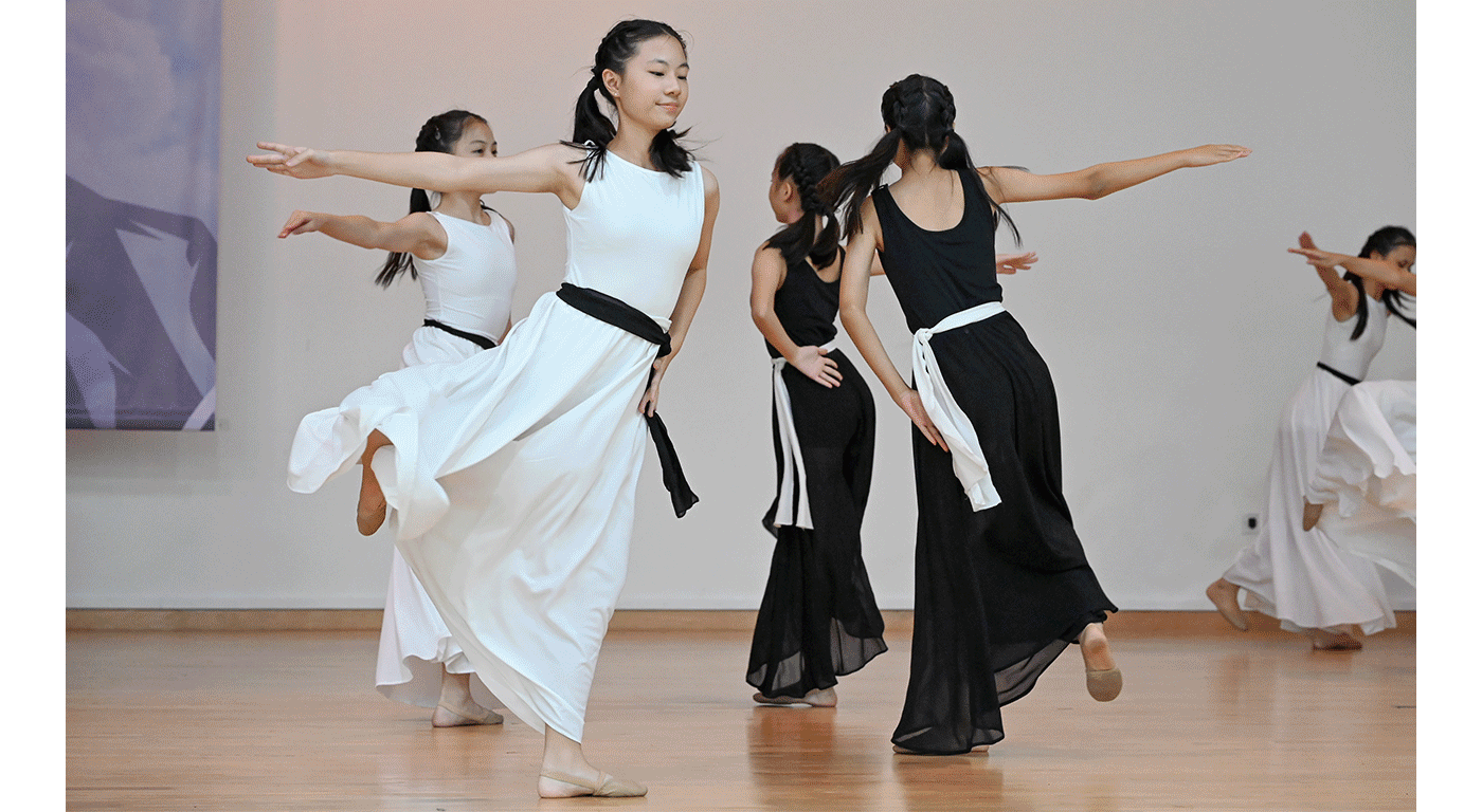 Young female dancers perform on a wood floor, wearing long black or white flowing dresses with contrasting sashes.