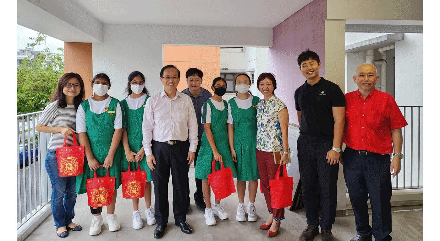 A group of ten people pose together, with four girls in green uniforms holding red Chinese New Year gift bags.