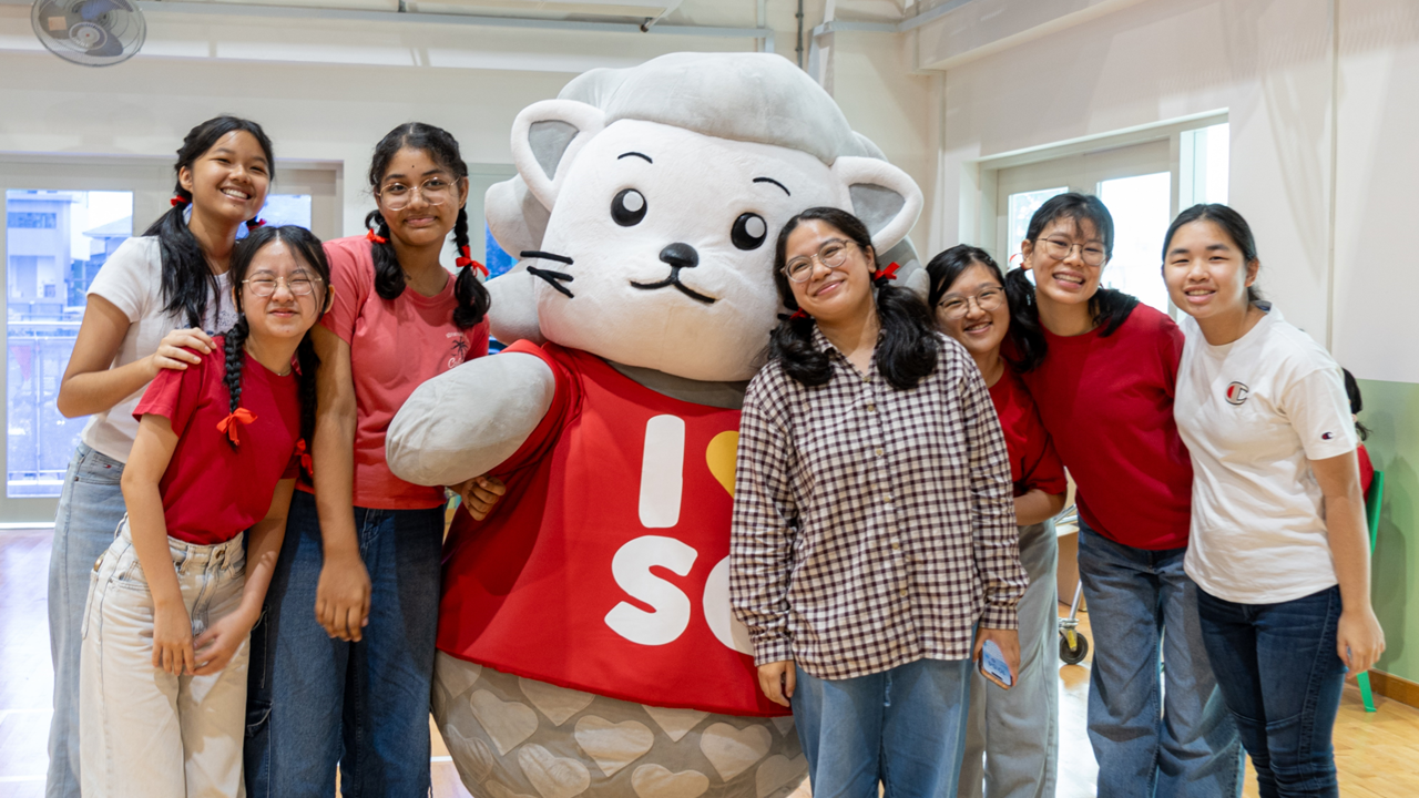 Seven smiling young women pose indoors with a large cat mascot wearing a red vest with text on it.