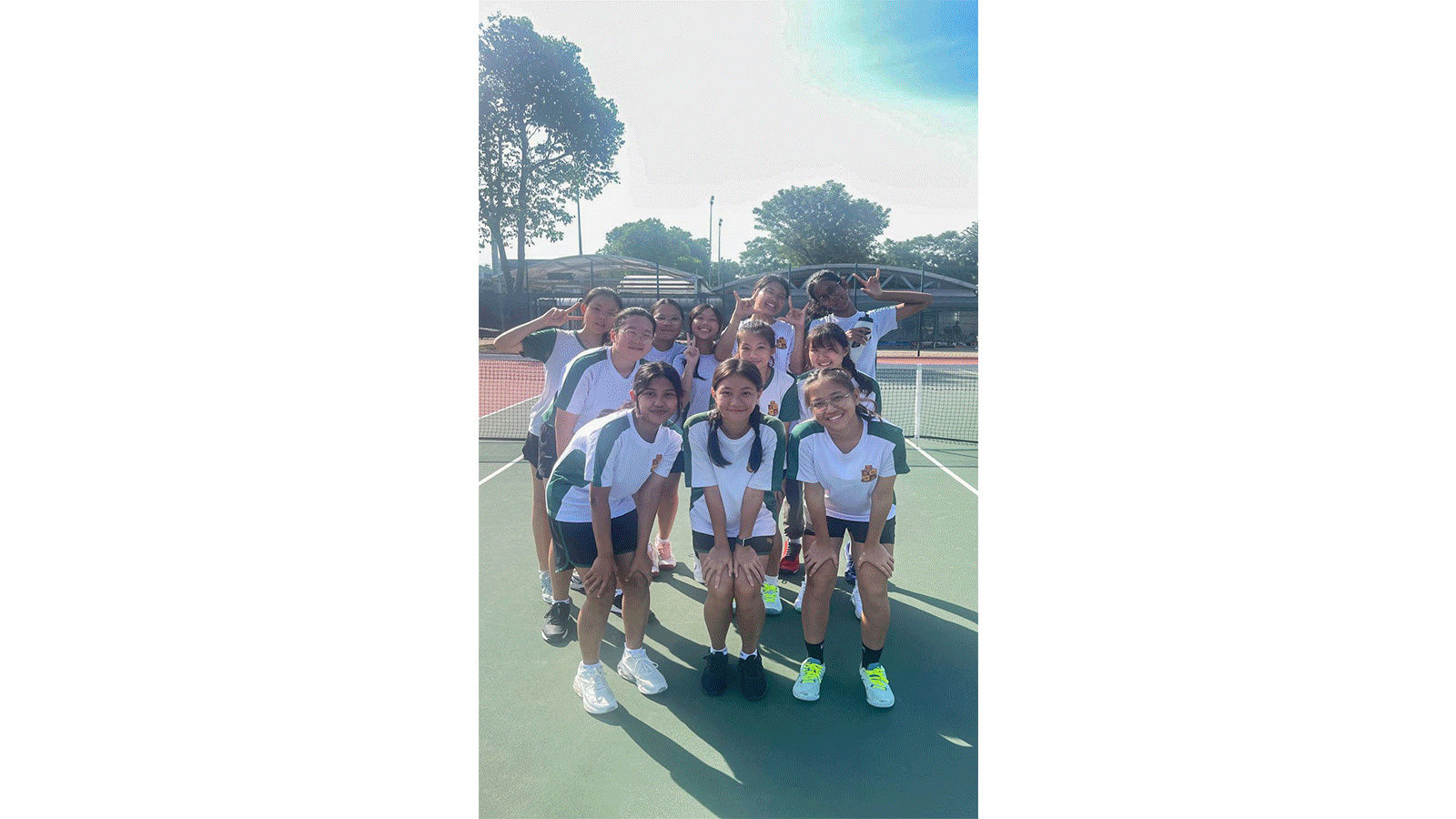 A group of young girls in matching white and green uniforms posing together on an outdoor tennis court.