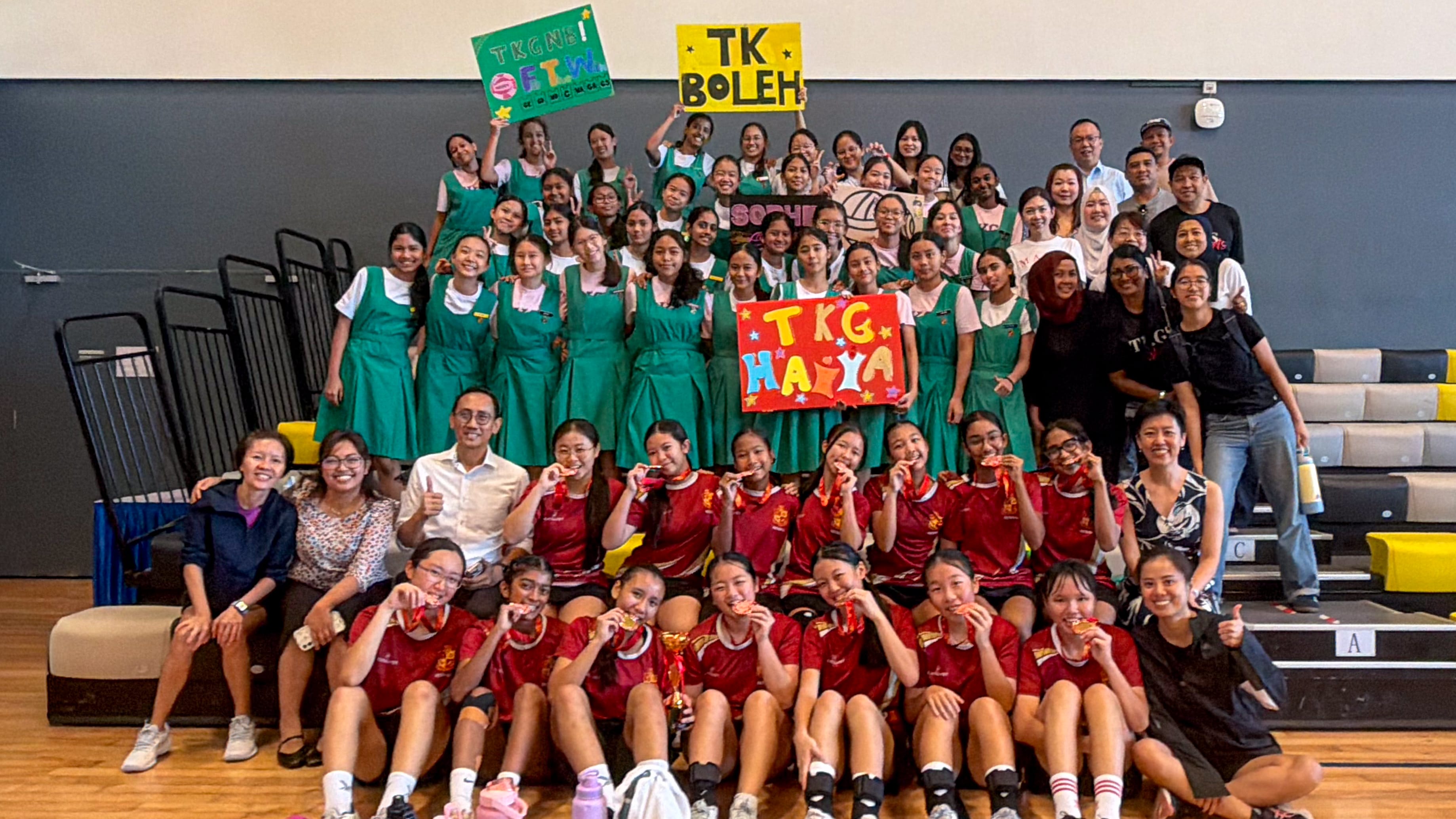 Large group in a gym; students in green uniforms and red jerseys pose with medals and TK signs.