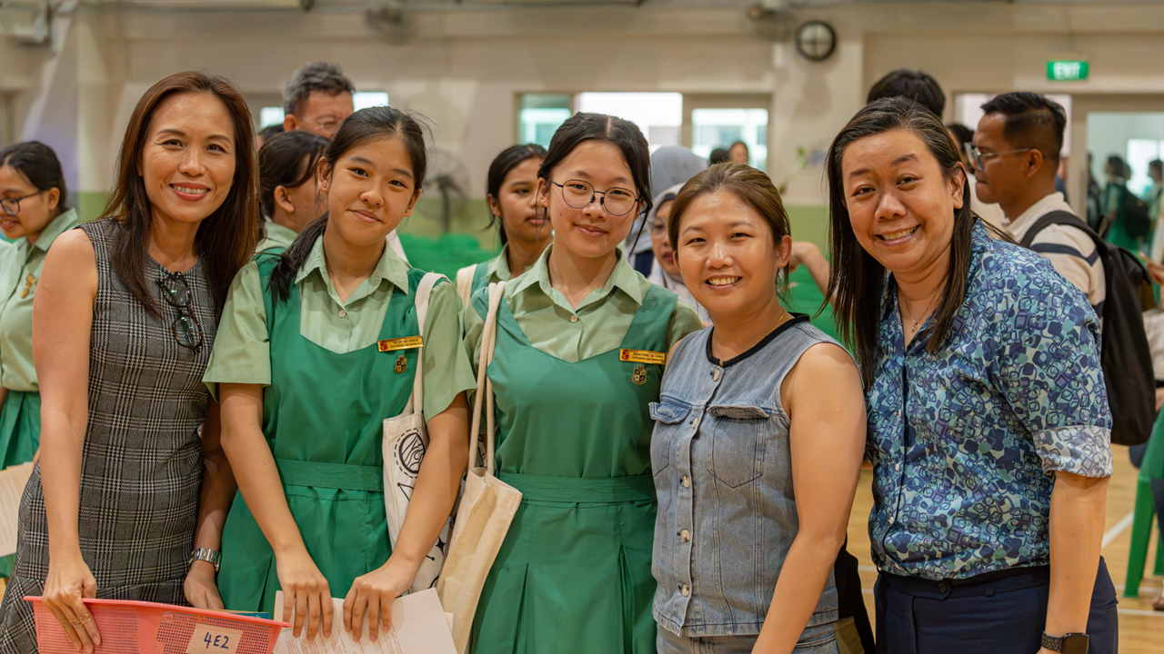 Five women smile at camera in a school hall, two wearing green school uniforms and three in casual adult clothing.