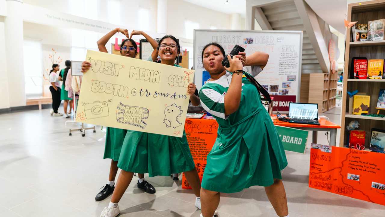Three students in green uniforms pose with a handmade "Visit Media Club Booth" sign and a camera in a library.
