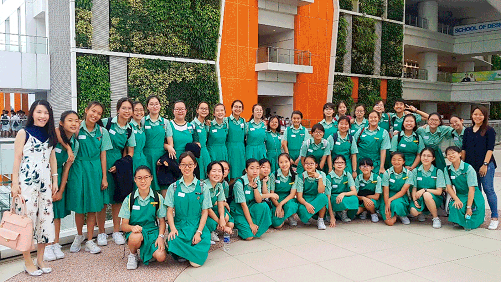 Group of students in green uniforms pose in a courtyard with a plant wall and orange building columns.