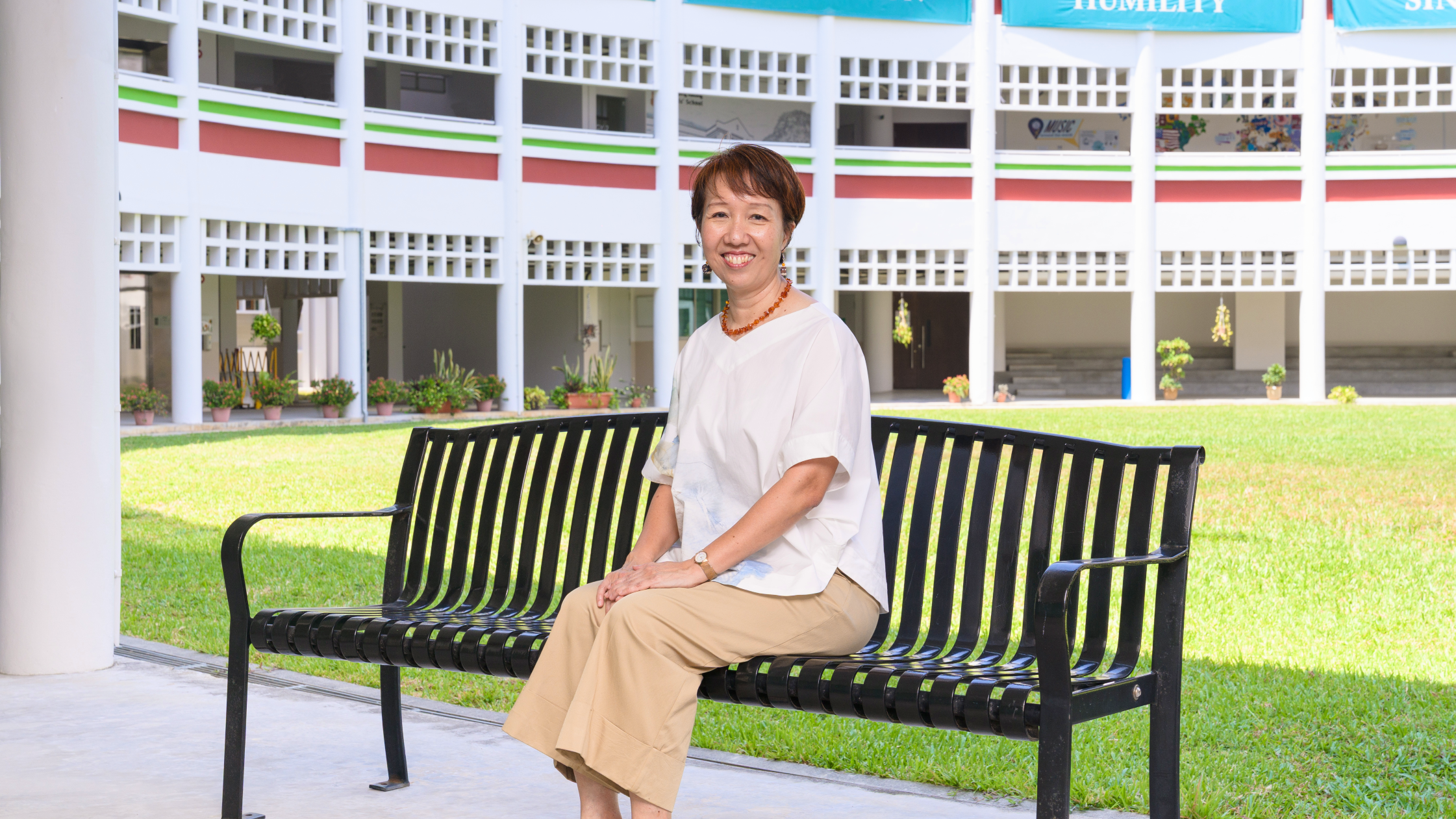 Smiling woman in a white top sits on a black bench in a grassy courtyard beside a white building.