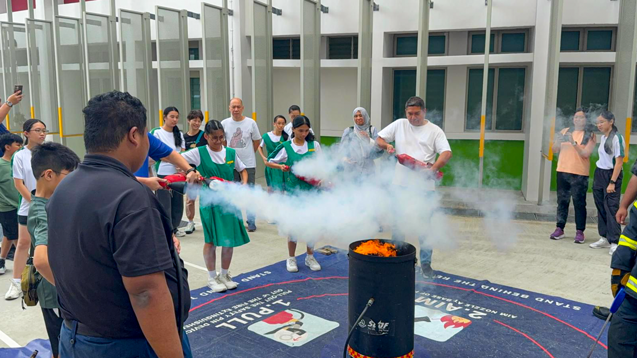 Students and adults use fire extinguishers on a burning barrel during an outdoor fire safety drill at a school.