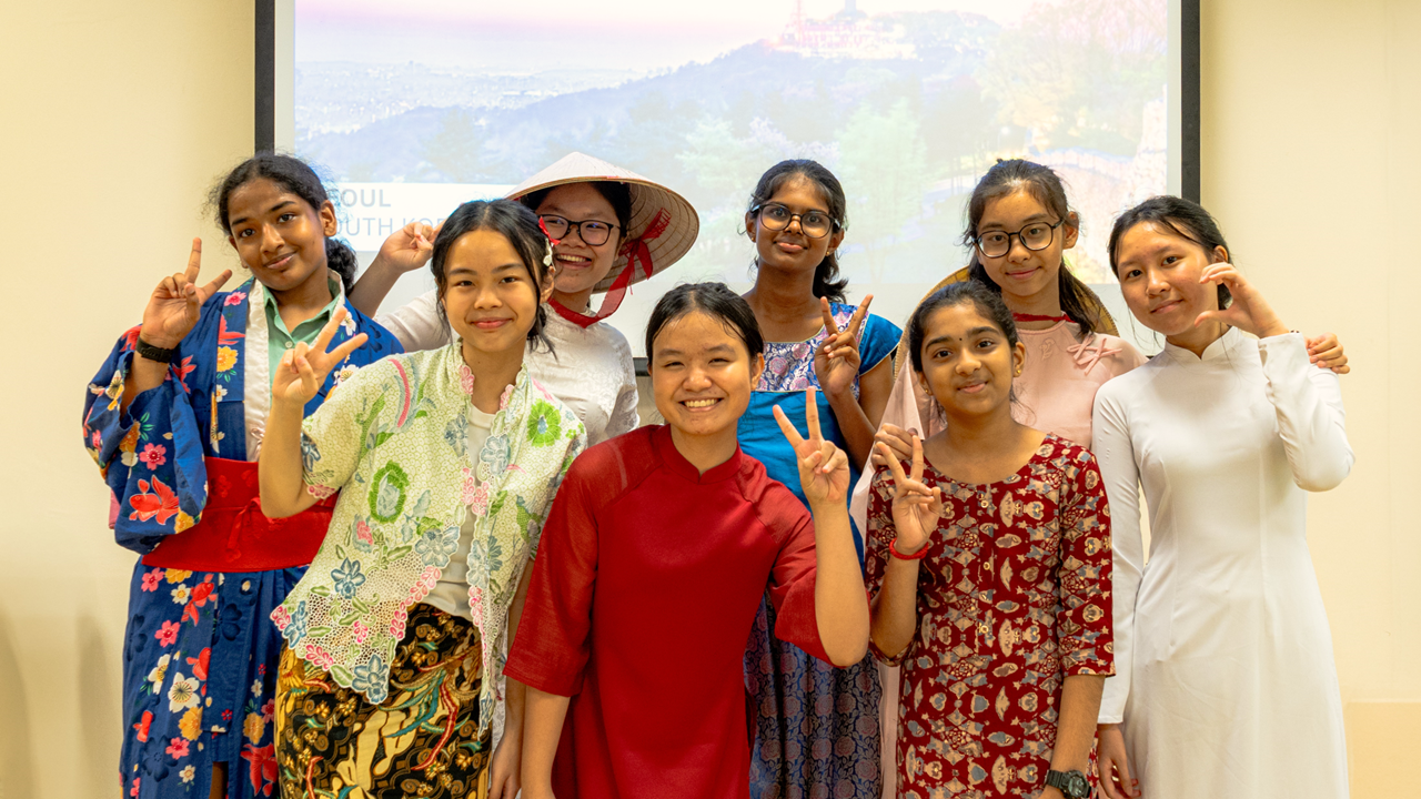Eight girls in traditional Asian attire pose smiling with peace signs in front of a projection screen.