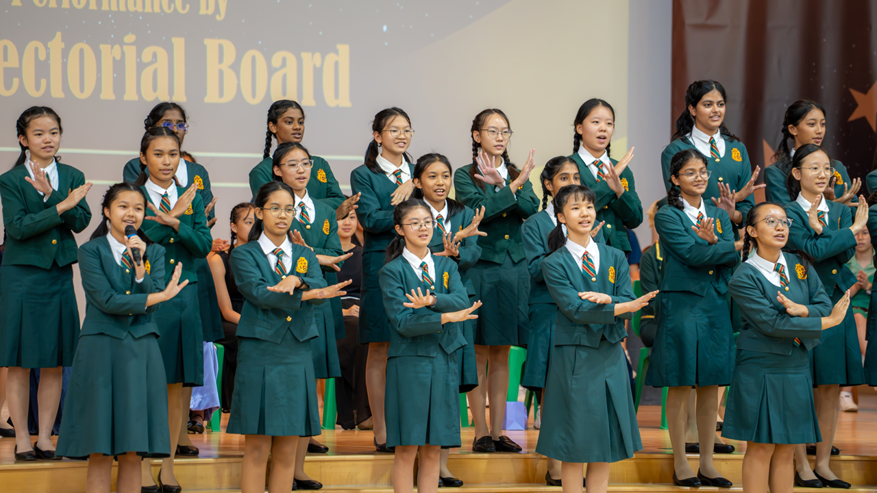 Girls in green school uniforms perform on stage, clapping and gesturing, with a projected screen reading "Rectorial Board."
