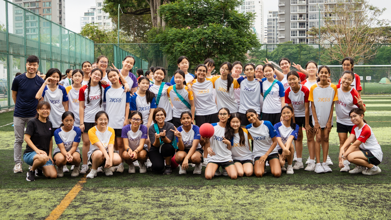 Large group of students in TKGS jerseys posing on a sports field, one holding a red ball, buildings in background.
