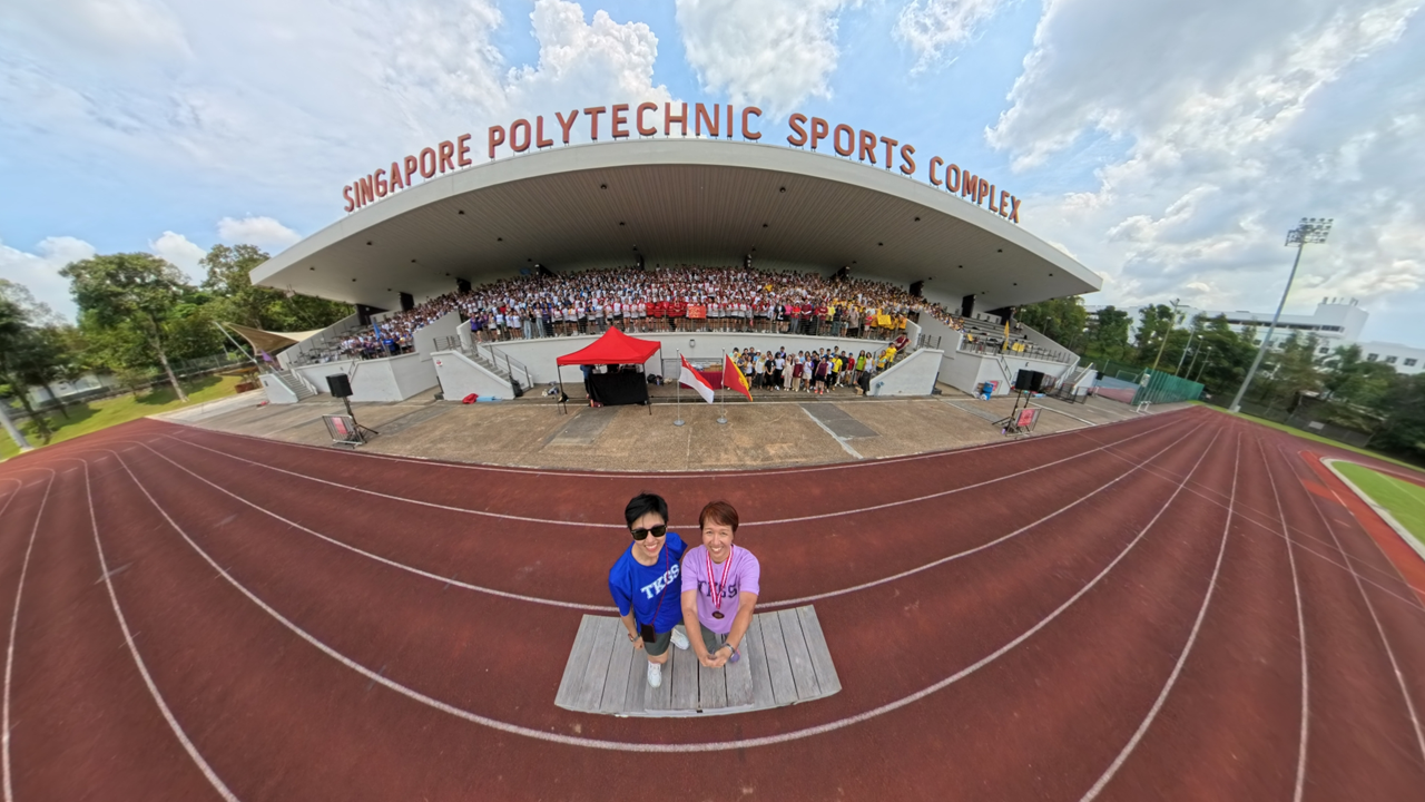Two people stand on a red running track at Singapore Polytechnic Sports Complex, with a large crowd in the grandstand behind them.