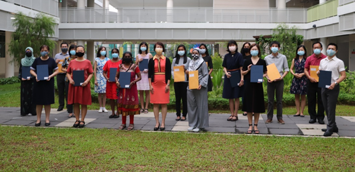 A group of masked people stand outdoors holding folders and envelopes, posing for a photo near a building.