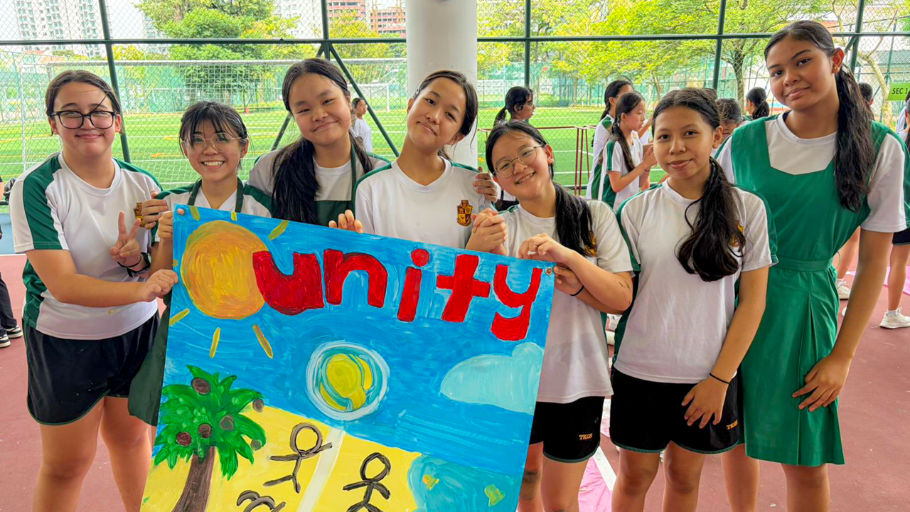 Seven schoolgirls in white and green uniforms smile while holding a painted poster reading Unity outdoors.