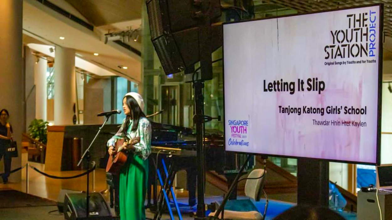 A young woman plays guitar at a microphone beside a screen reading "Letting It Slip, Tanjong Katong Girls' School."