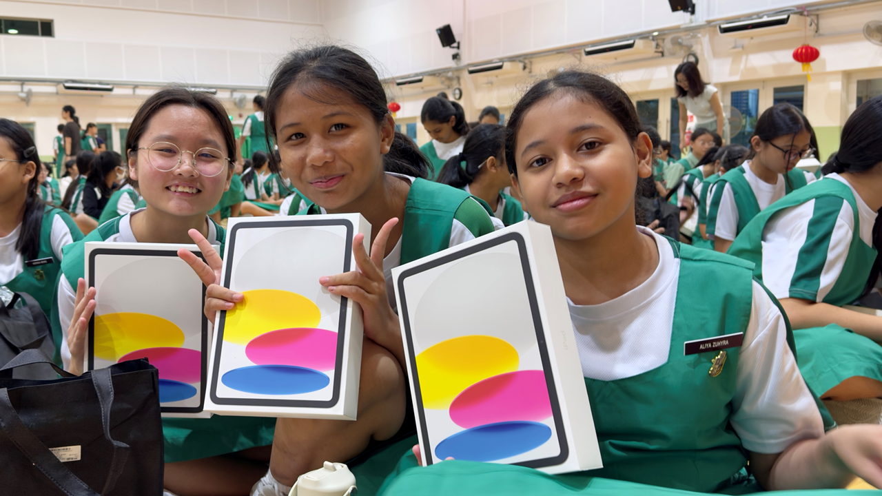 Three schoolgirls in green uniforms smile while holding Apple iPad boxes in a school hall.