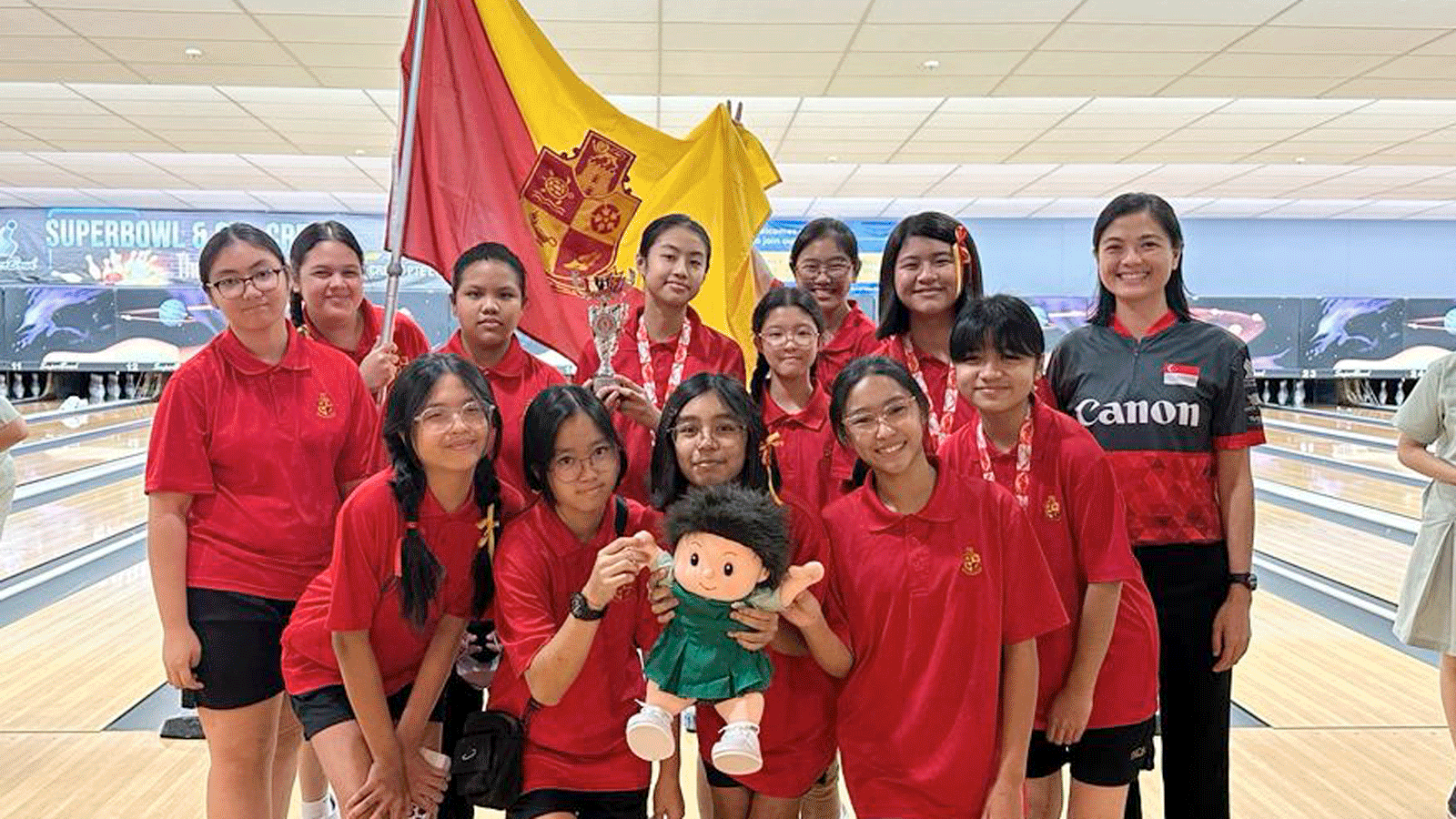 A group of girls in red uniforms pose at a bowling alley holding a trophy, a flag, and a small doll.