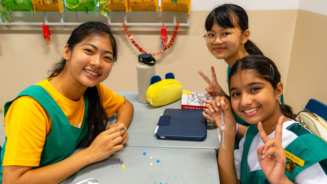 Three smiling schoolgirls in green uniforms sit at a desk with beads, a tablet, and a water bottle.