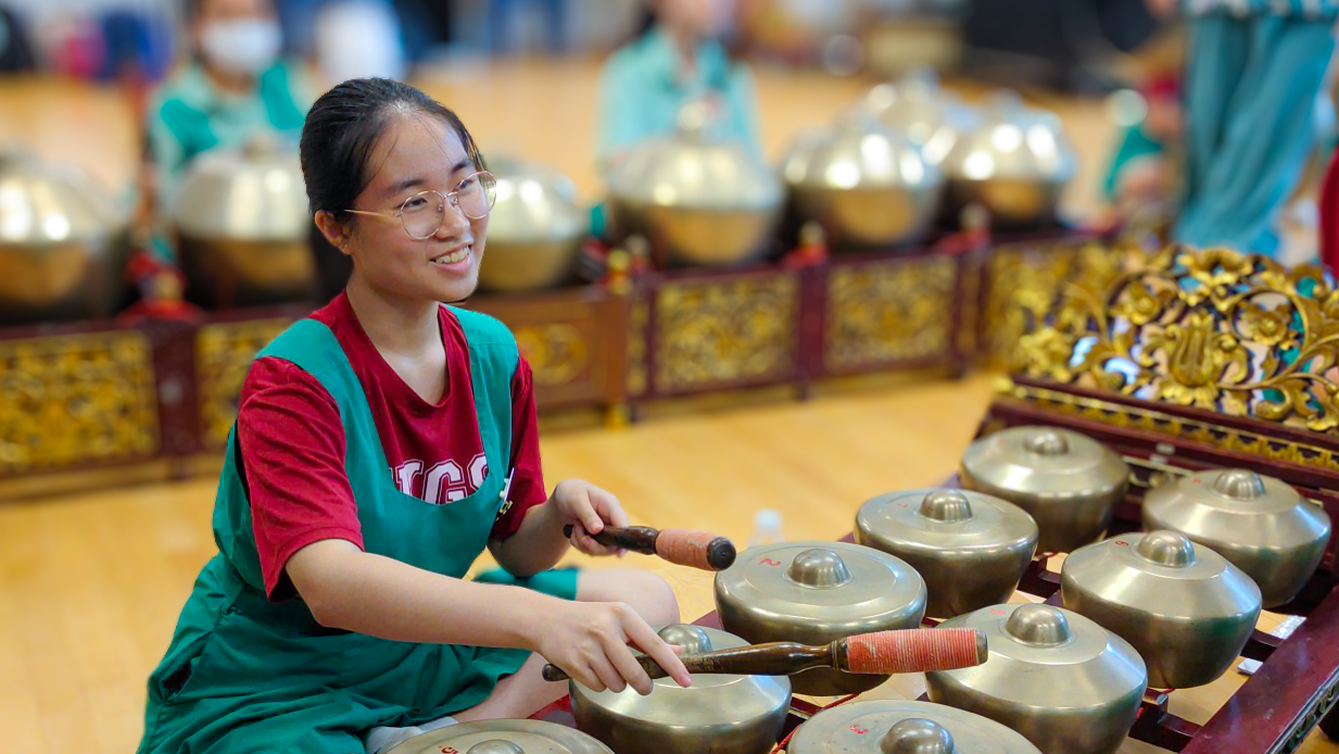 Smiling young woman with glasses playing a gamelan, striking bronze gong-chime instruments with mallets.