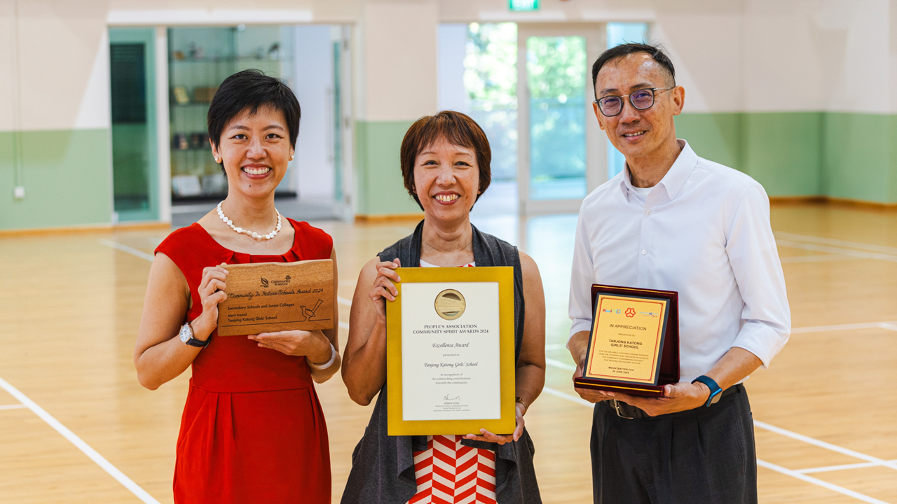 Three smiling people stand in a gymnasium holding award plaques and a framed certificate for Tanjong Katong Girls School.