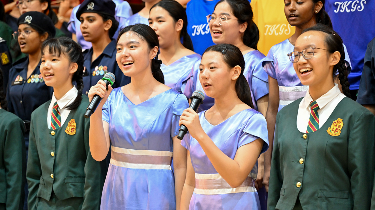 Students in school uniforms and blue dresses sing on stage, two holding microphones, with a large group behind them.