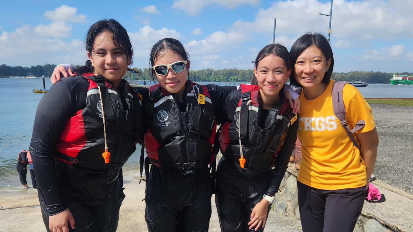 Four smiling women pose by a waterfront, three wearing black life vests and one in a yellow TKGS shirt.
