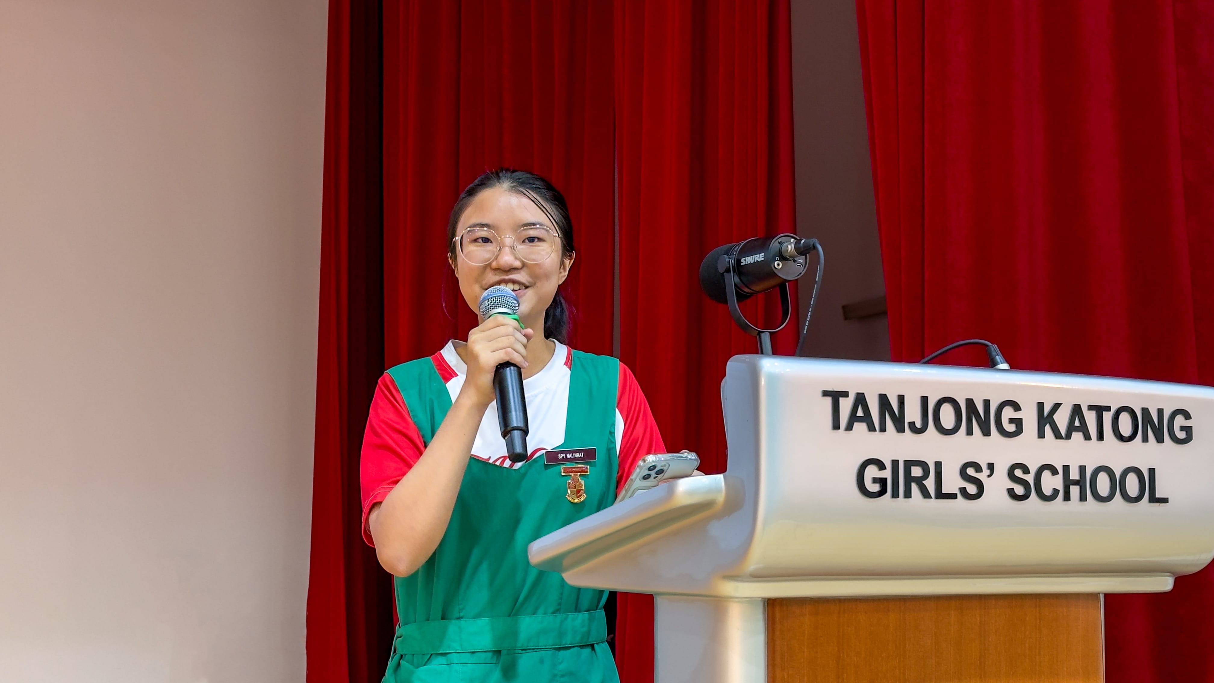 A young girl in a green and red uniform speaks into a microphone at a Tanjong Katong Girls' School podium.
