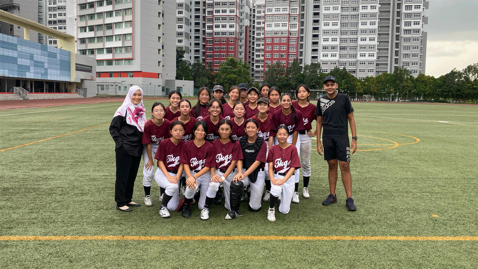 A girls softball team in maroon uniforms poses with two coaches on a green field, with tall buildings behind them.