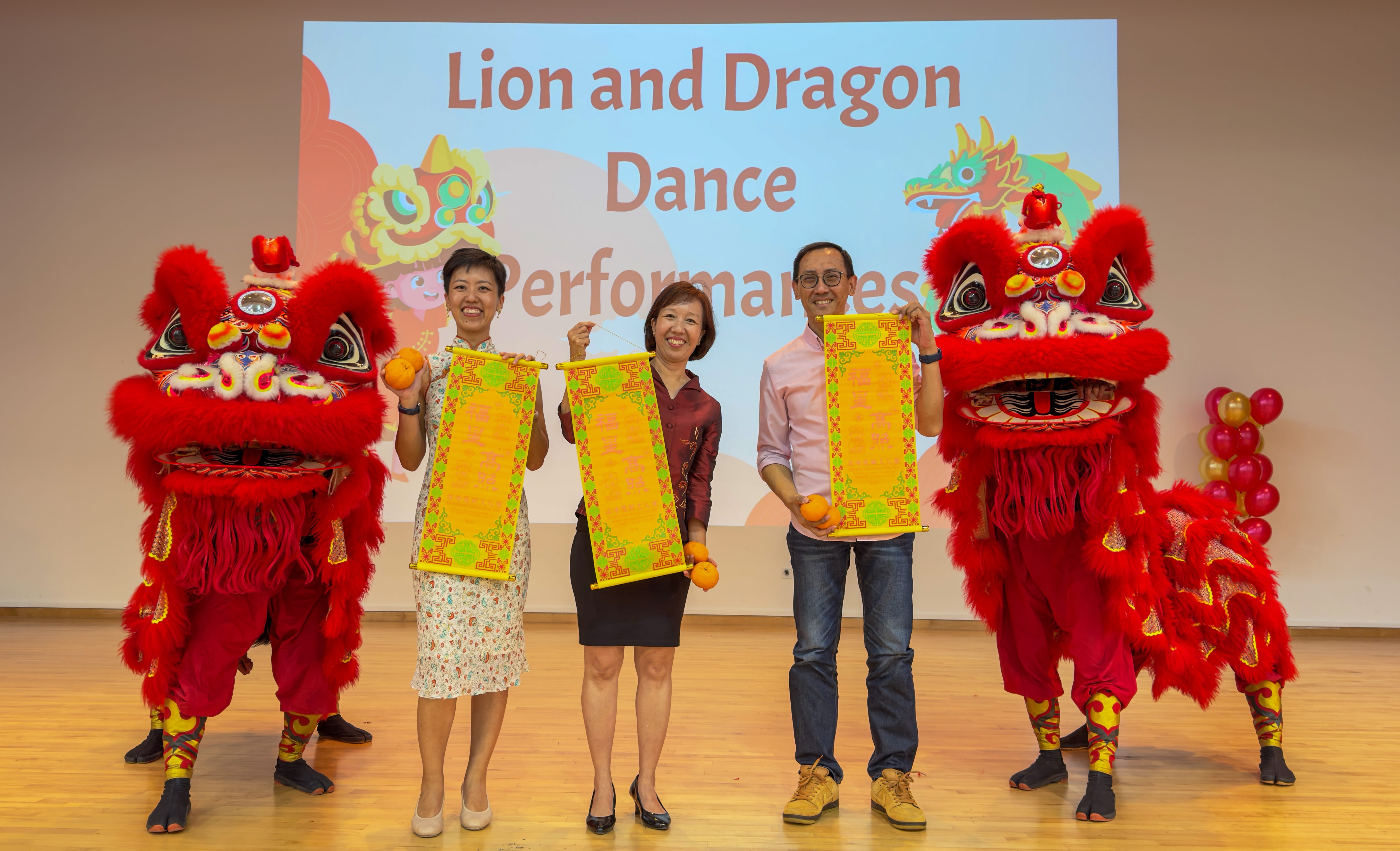 Three people hold decorative scrolls and oranges flanked by two red lion dance costumes on a stage.