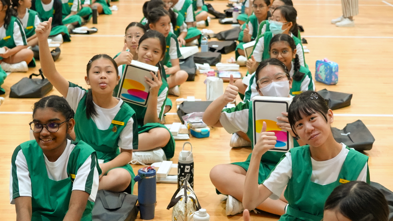 Girls in green and white school uniforms sit on a gym floor, giving thumbs up while holding new tablet boxes.