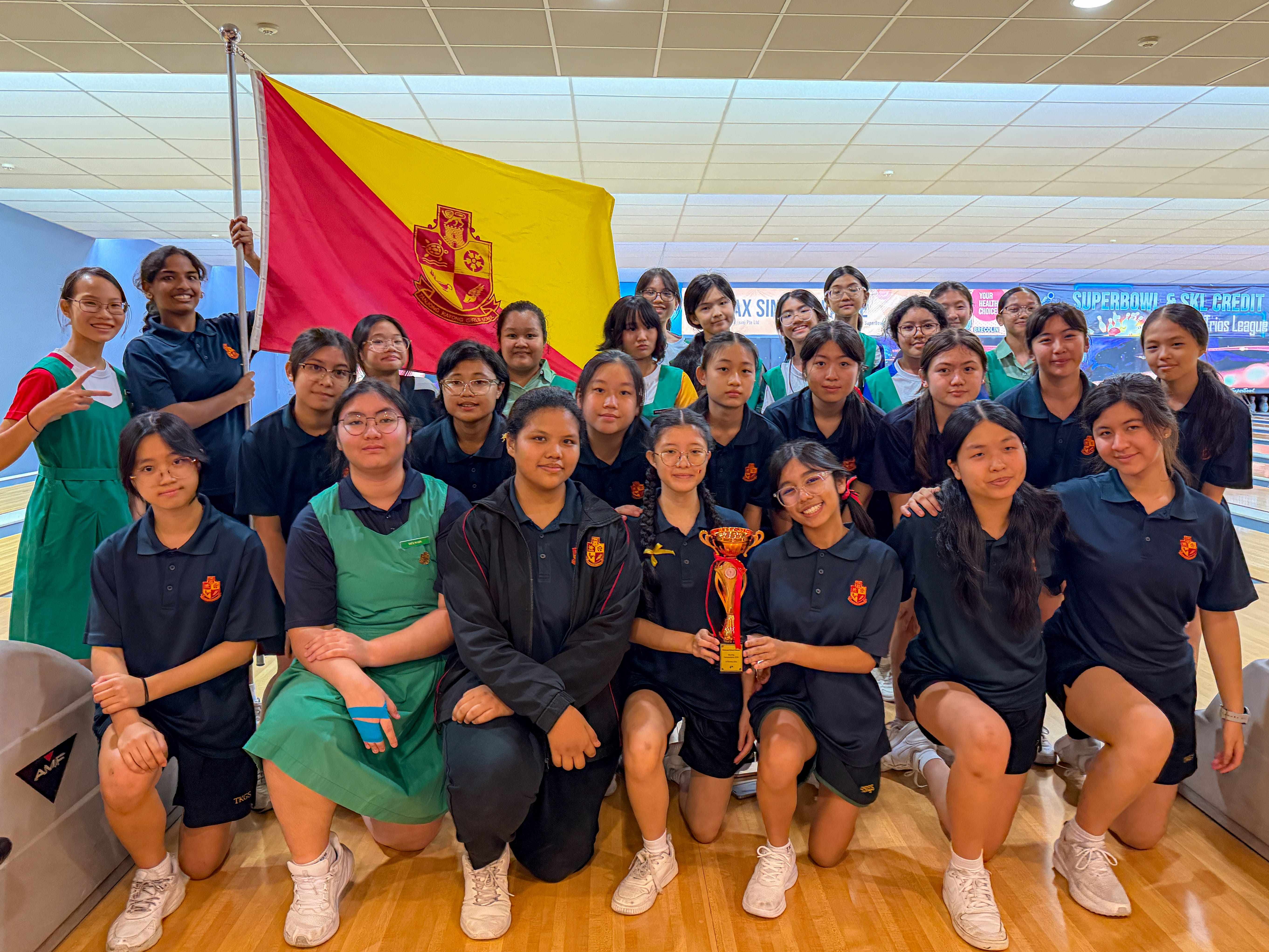 A group of young female students pose with a trophy and a red and yellow school flag at a bowling alley.