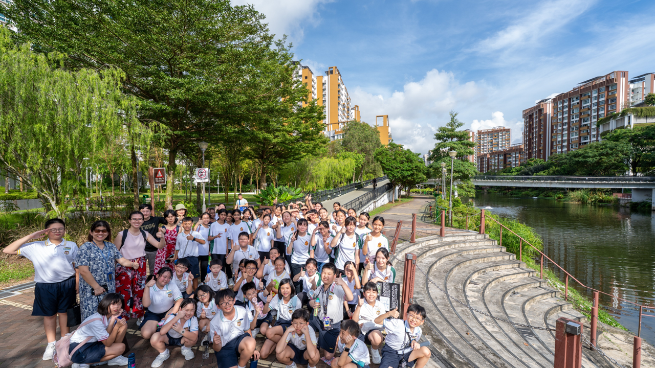 A large group of students in white uniforms poses near a canal path with trees and tall buildings in the background.