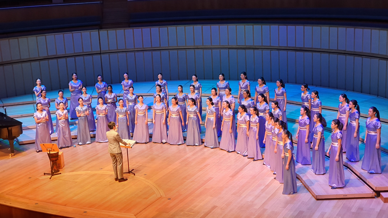 A conductor leads a large women's choir in purple gowns on a concert hall stage with blue lighting.