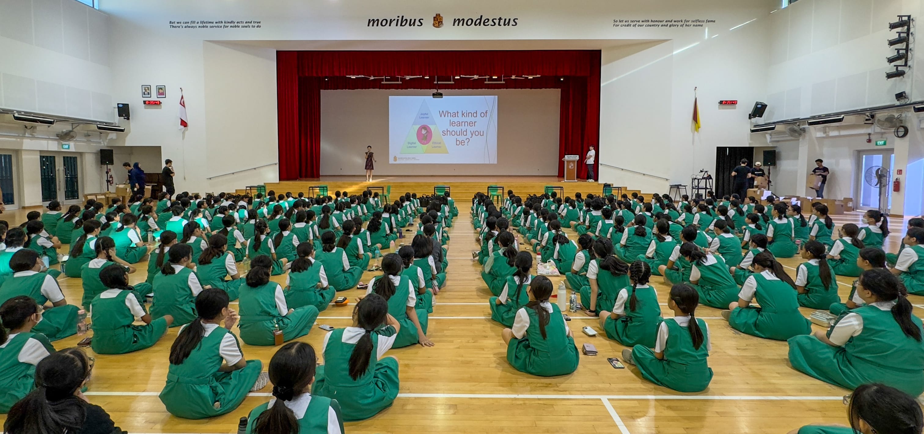 Students in green uniforms sit on a school hall floor facing a stage with a projected slide reading "What kind of learner should you be?"