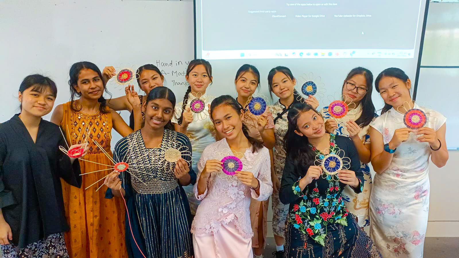 A group of smiling girls in traditional outfits hold colorful woven flower crafts in a classroom.