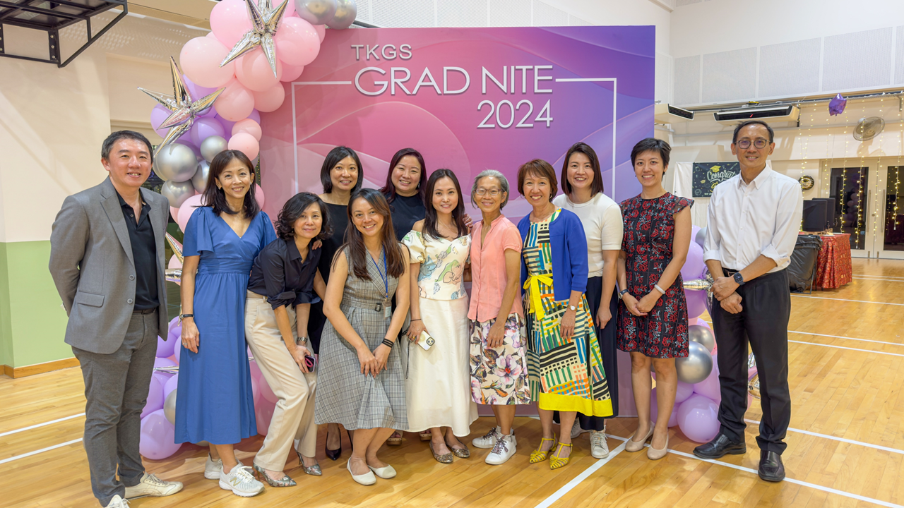 Group of adults posing in front of a TKGS Grad Nite 2024 banner with pink and purple balloon decorations.