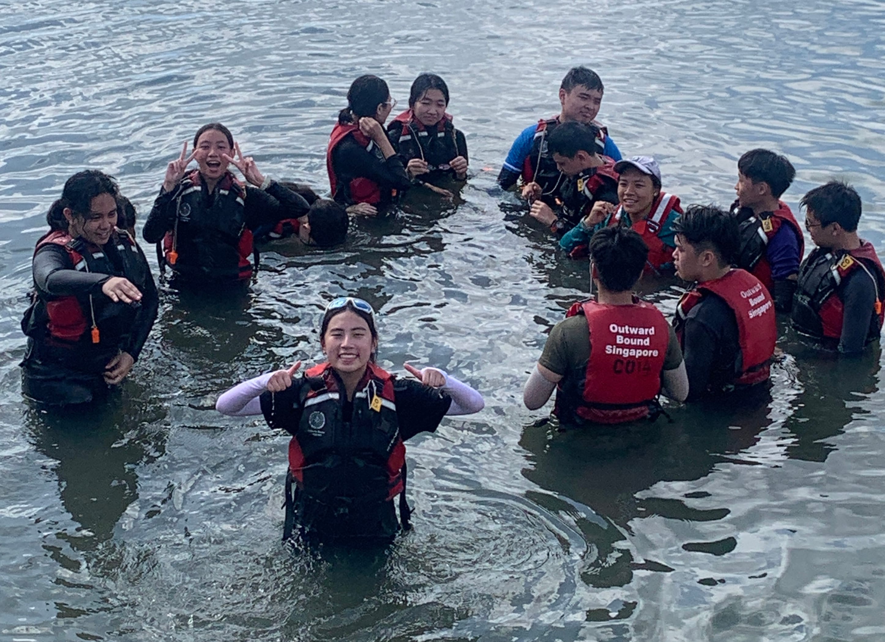 A group of smiling young people wearing red Outward Bound Singapore life vests stand waist-deep in open water.