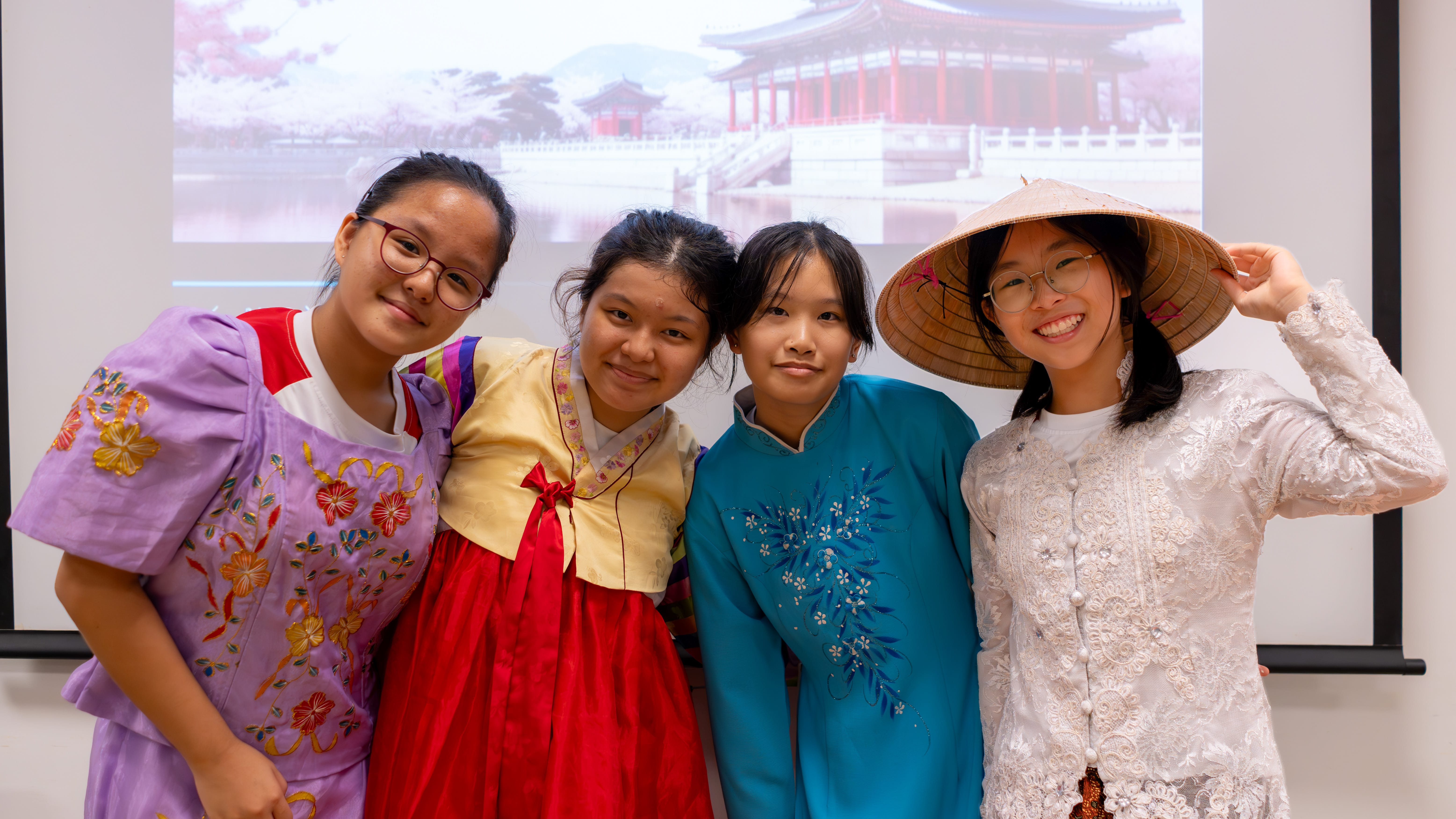 Four girls in colorful traditional outfits pose smiling; one wears a conical hat, a temple photo behind them.